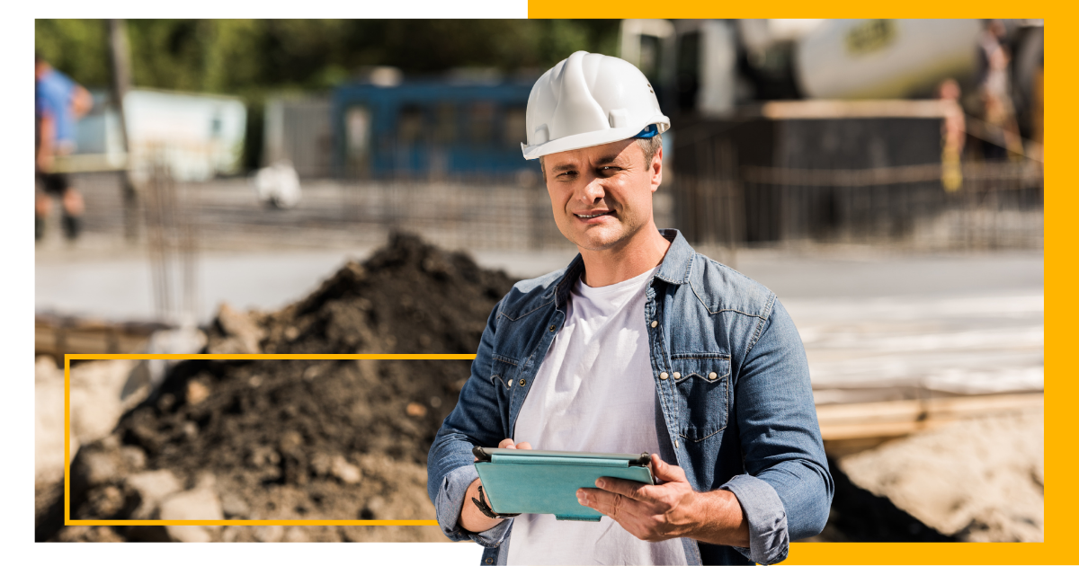 Man stands in a construction site holding a tablet with a white hard hat on